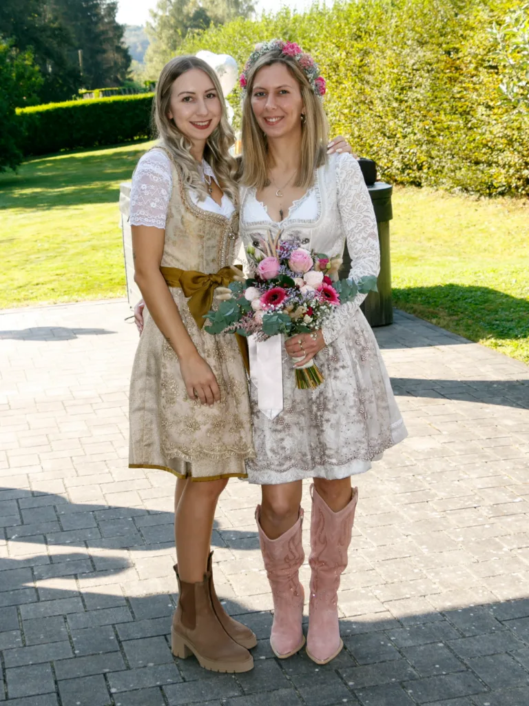 Braut im weißen Spitzen-Dirndl mit Blumenkranz neben ihrer Begleitung im goldenen Dirndl